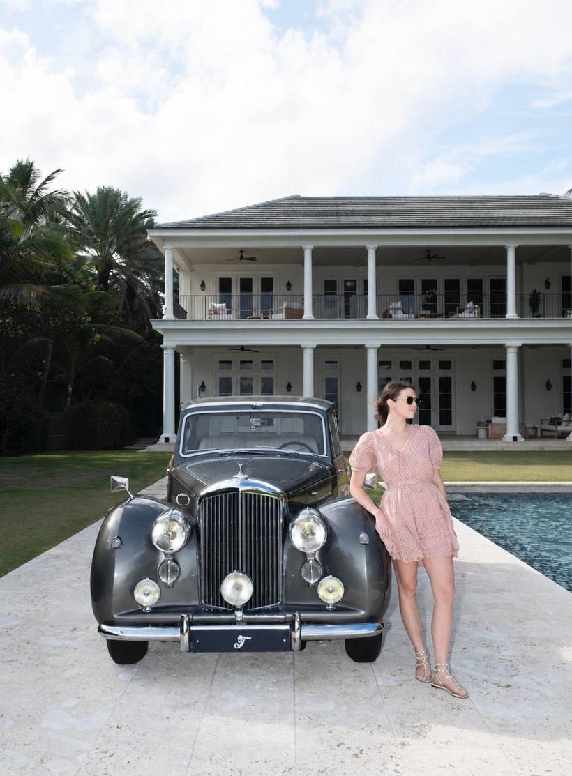 Pink dress, leaning on a vintage car in front of an estate